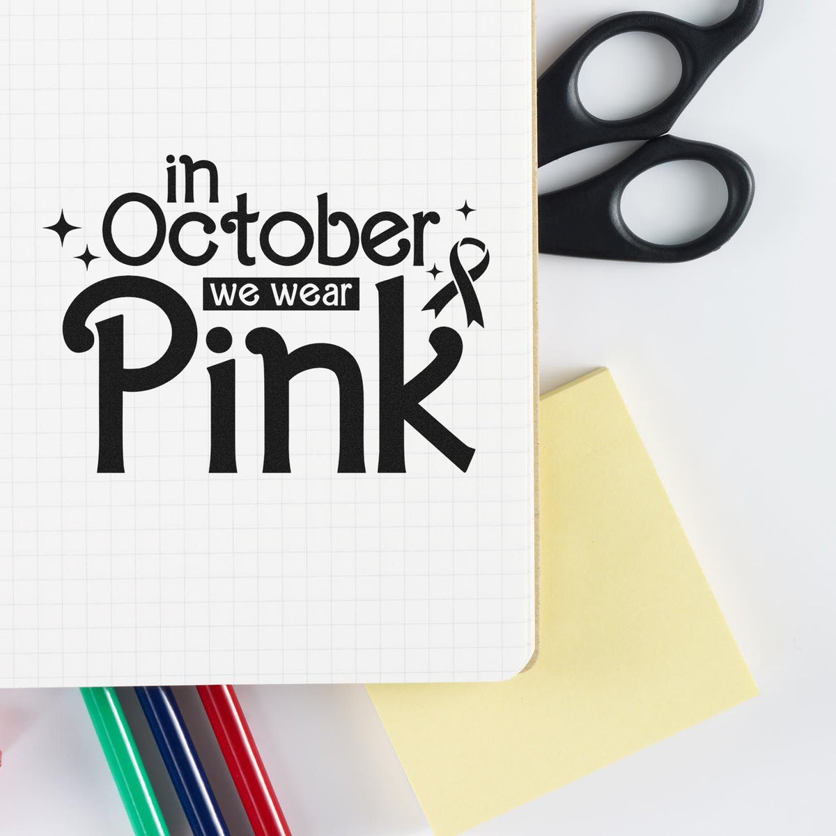 Black stamp and seal design reading in October we wear Pink on white grid paper, with scissors, yellow note, and colored pens.