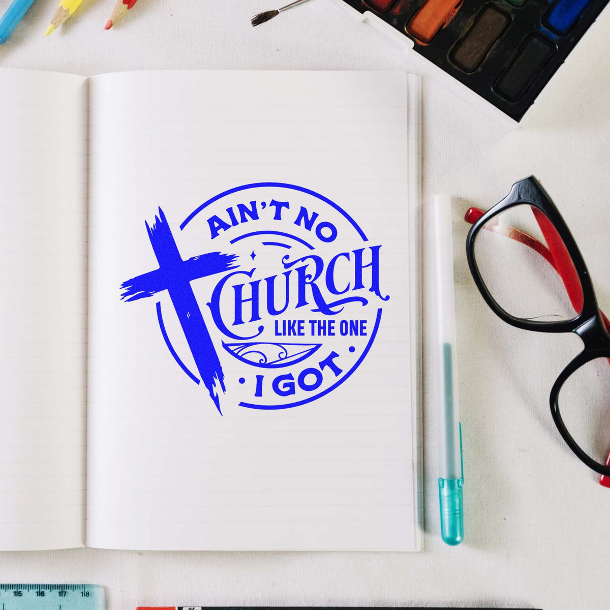 Blue church stamp seal reading Aint no church like the one I got on an open notebook, with glasses and watercolor supplies.