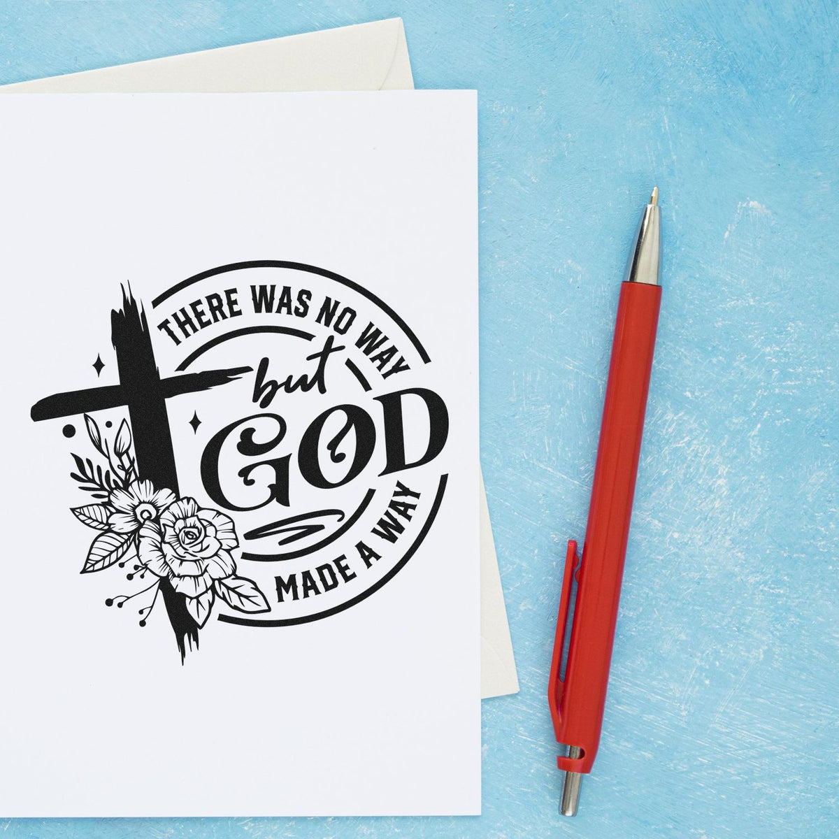 Black faith rubber stamp seal reading There was no way but God with floral cross on white card, beside a red pen on blue background.