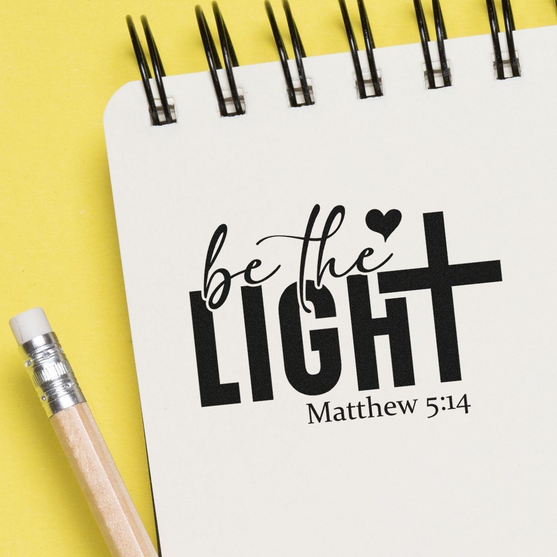 Christian stamp and seal reading be the light Matthew 5:14 on a white spiral notebook over a yellow background, with a pencil.
