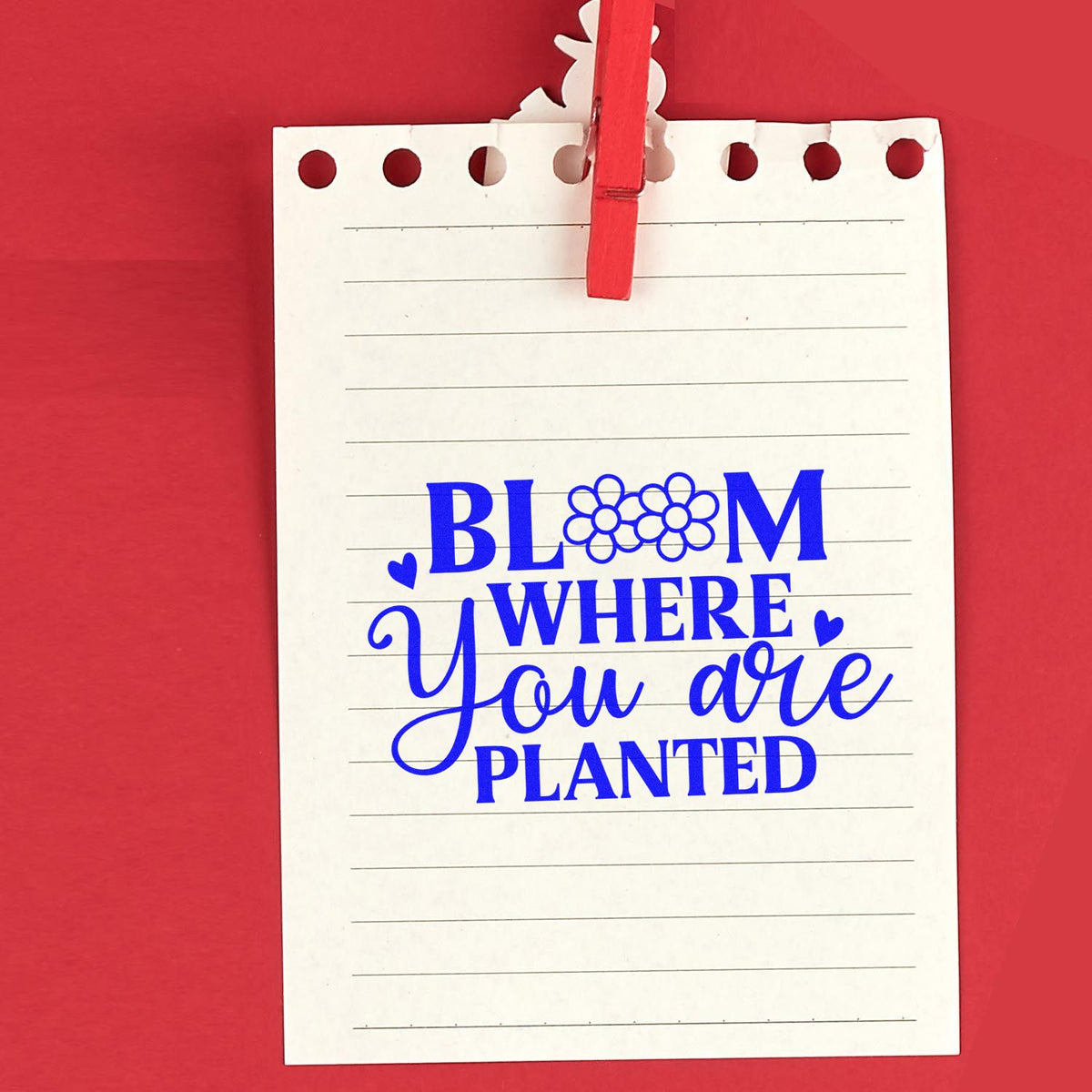 Blue floral stamp design on white lined notepad, clipped with a red clothespin, reading Bloom where you are planted on red background.