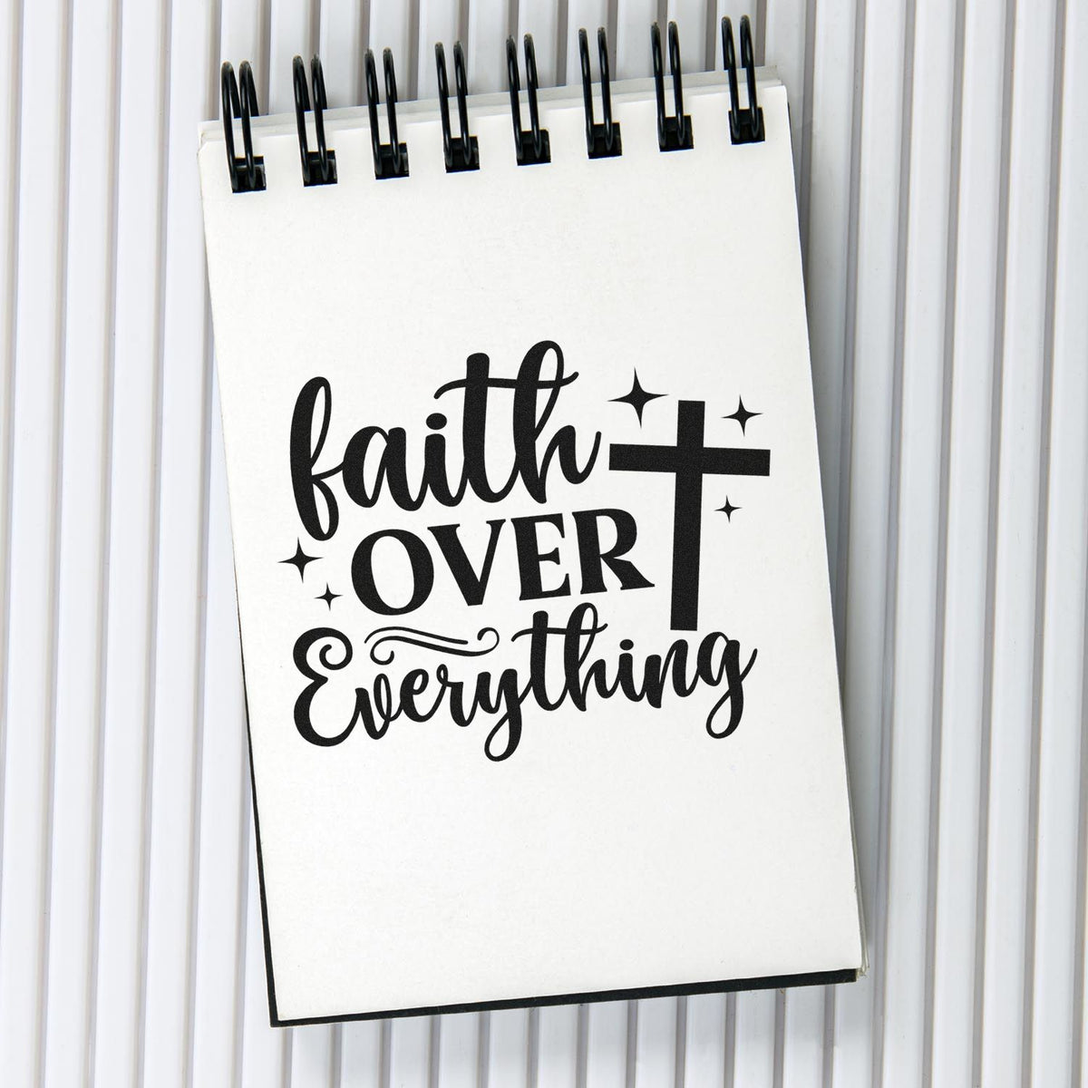 Black faith over everything stamp design on a white spiral notepad with cross and star accents