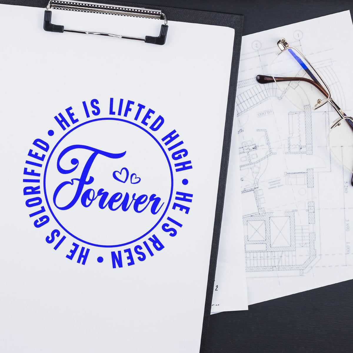 Blue custom stamp and seal reading He is lifted high, He is risen, glorified forever on clipboard beside glasses and plans.