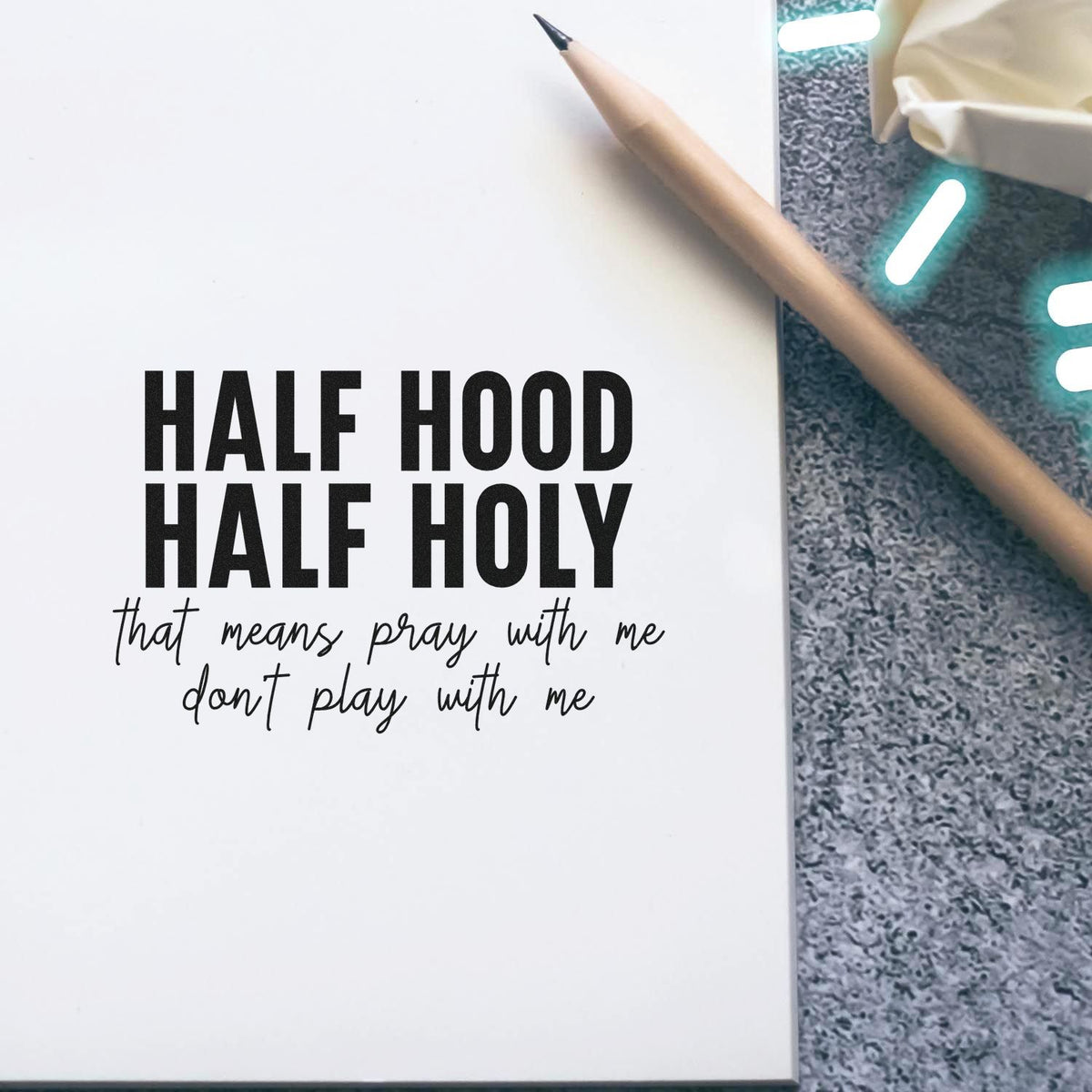 Black custom stamp seal on white paper reading HALF HOOD HALF HOLY with handwritten slogan, beside a tan pencil on gray surface.