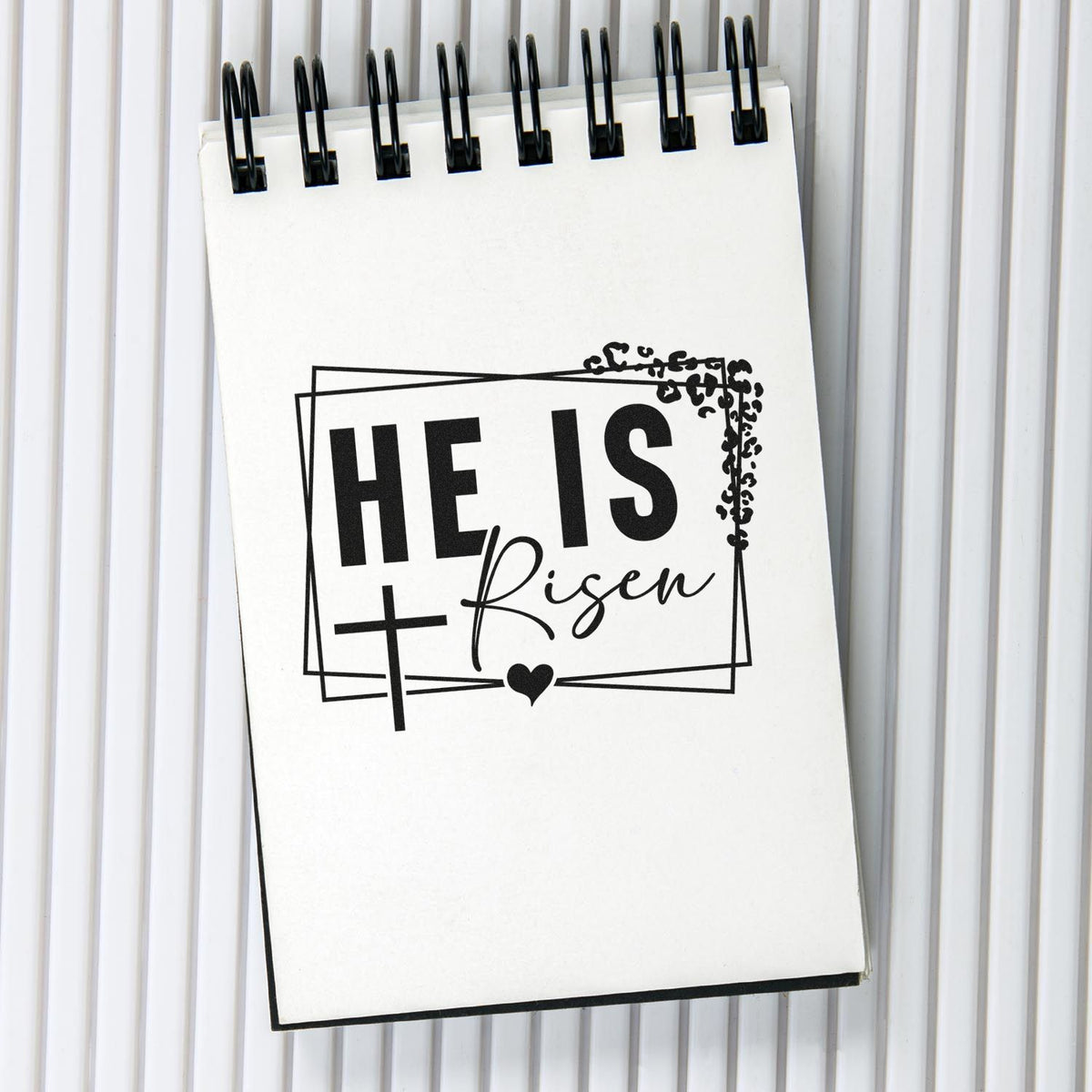 Black Christian stamp seal reading He Is Risen with cross, heart, on a white spiral notepad