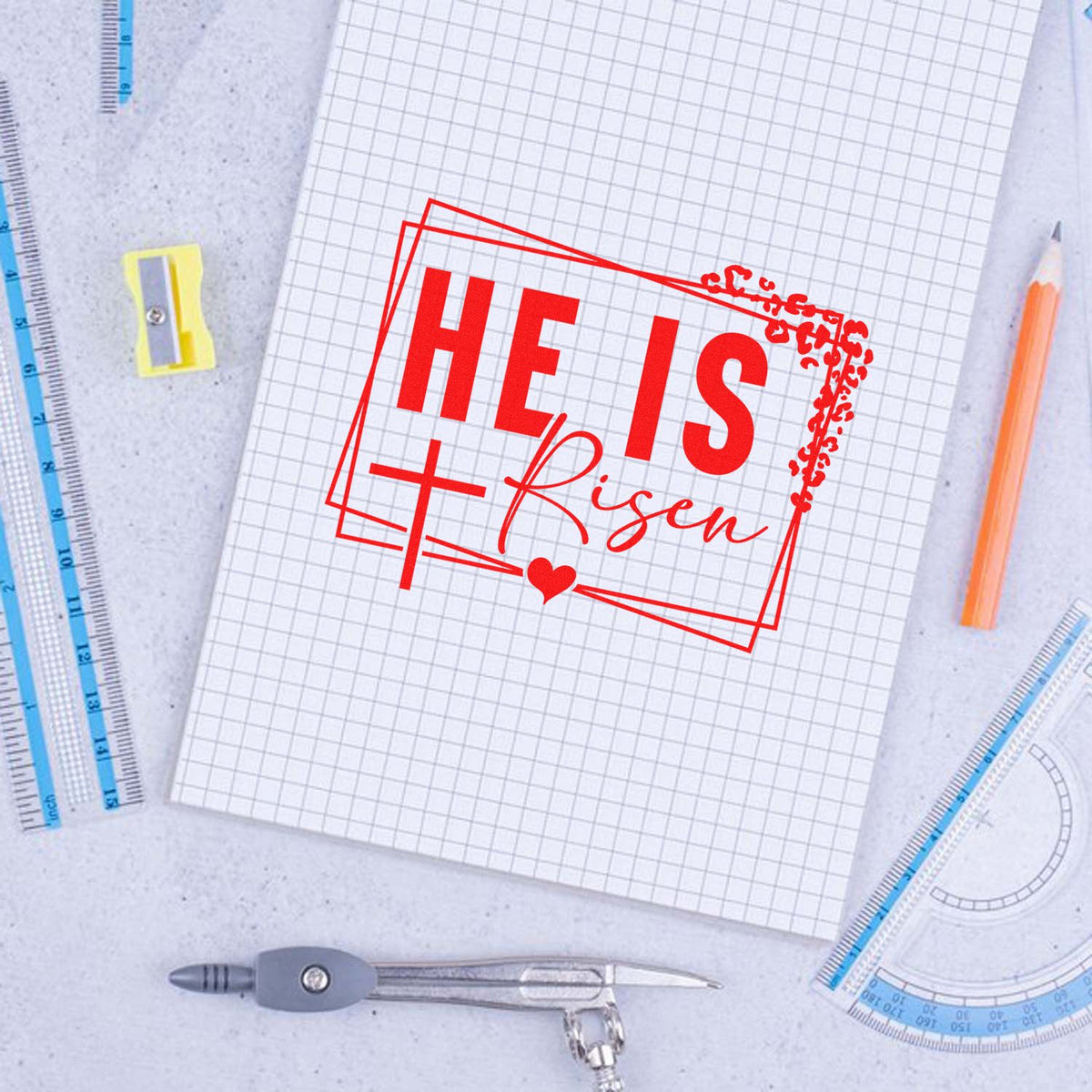 Red HE IS Risen stamp and seal on graph paper notebook, with blue rulers, orange pencil, sharpener, and compass.