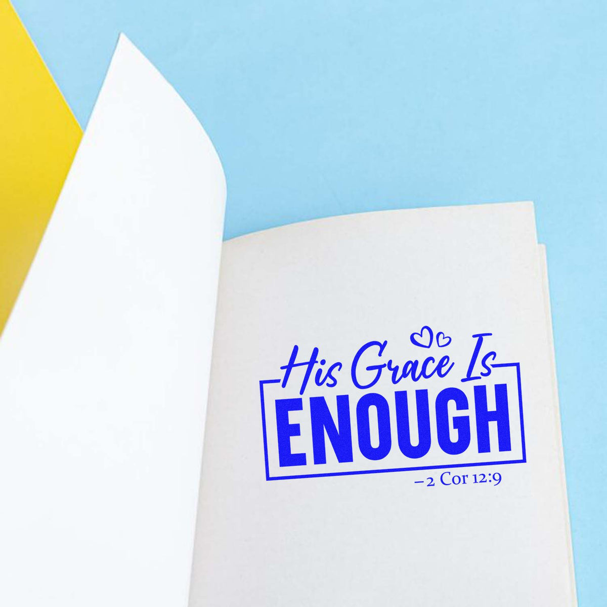 Blue stamp and seal reading His Grace Is ENOUGH with hearts and 2 Cor 12:9 on white paper over a light blue background.