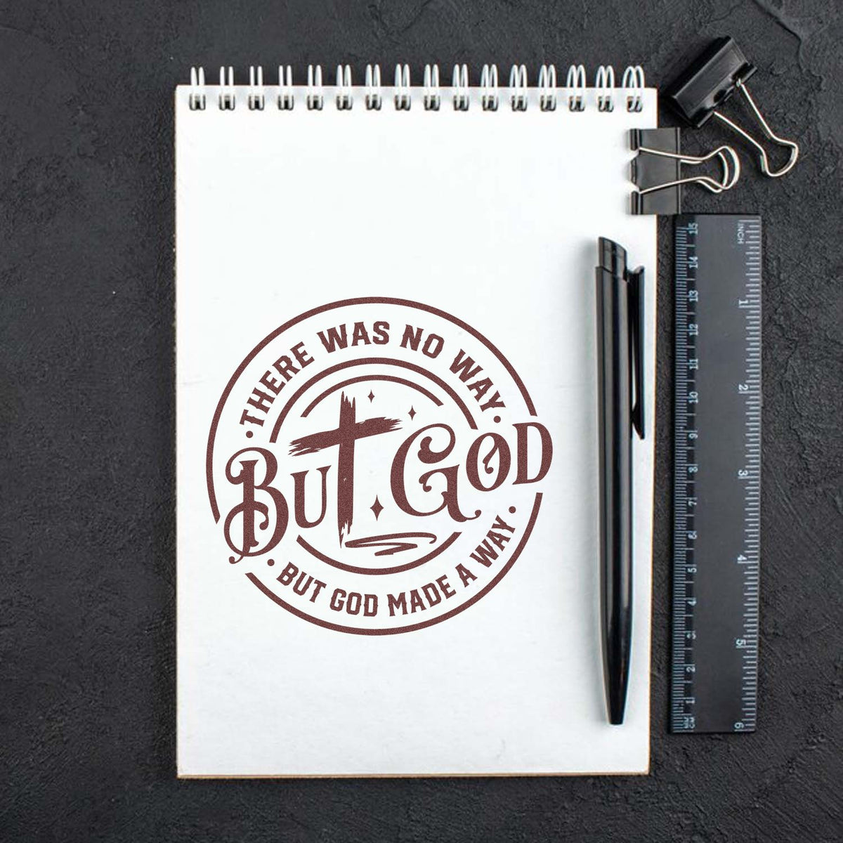 Brown circular rubber stamp seal reading But God on a white spiral notebook, with black pen, ruler, and binder clips on a dark background.