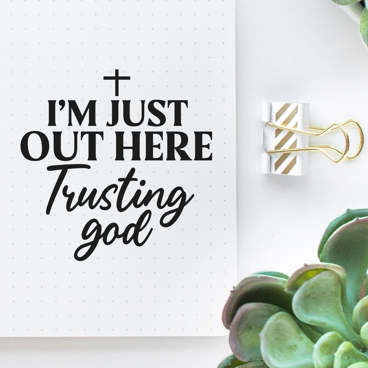 Black faith stamp reading Im just out here trusting God on white dotted paper with gold striped binder clip and green succulent.