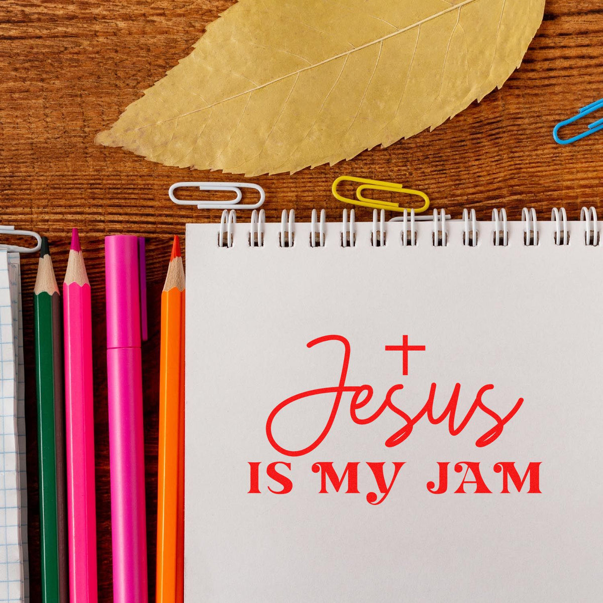 Red Jesus is my Jam stamp on a white spiral notebook, with colorful pencils, paper clips, and a tan leaf.