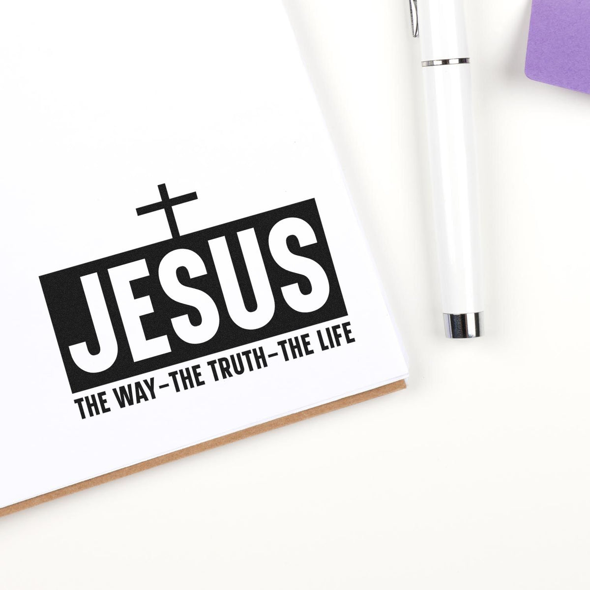 Black JESUS seal stamp with cross and tagline on white paper, beside a white pen and purple card.