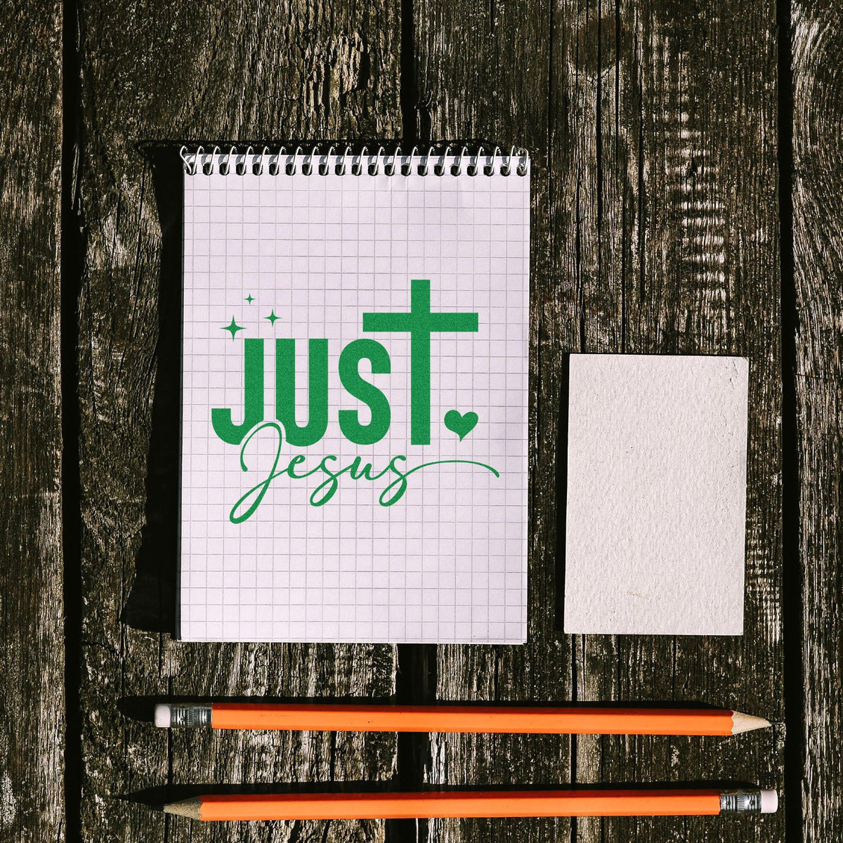 Green Just Jesus stamp and seal set on graph notebook, with blank white card and two orange pencils on dark wood.