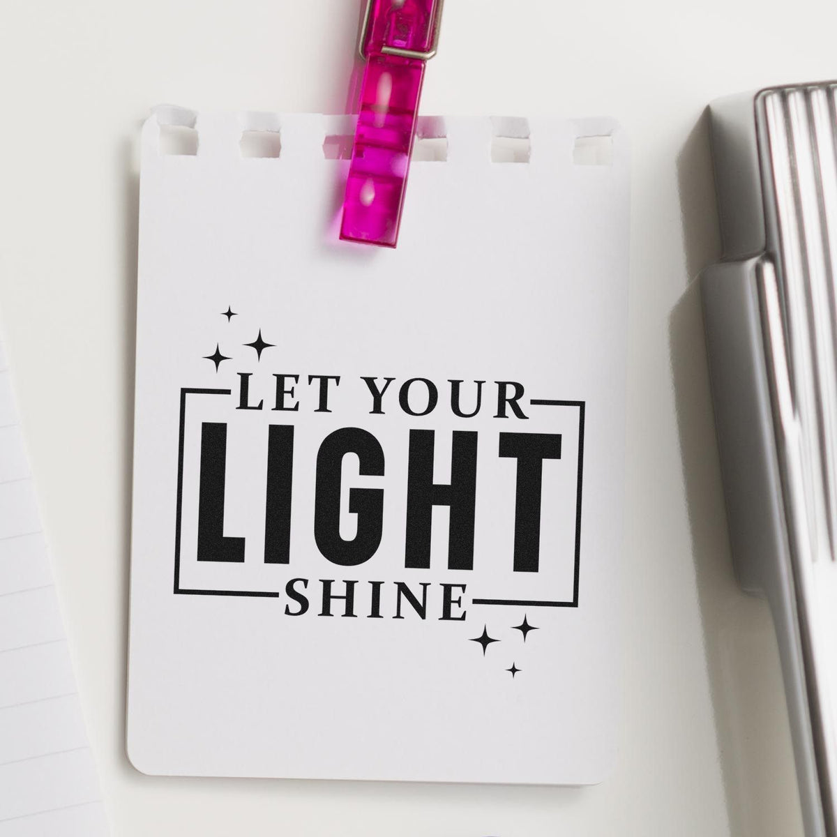 White stamp card clipped with pink seal clip, featuring black Let Your Light Shine text and sparkle accents.