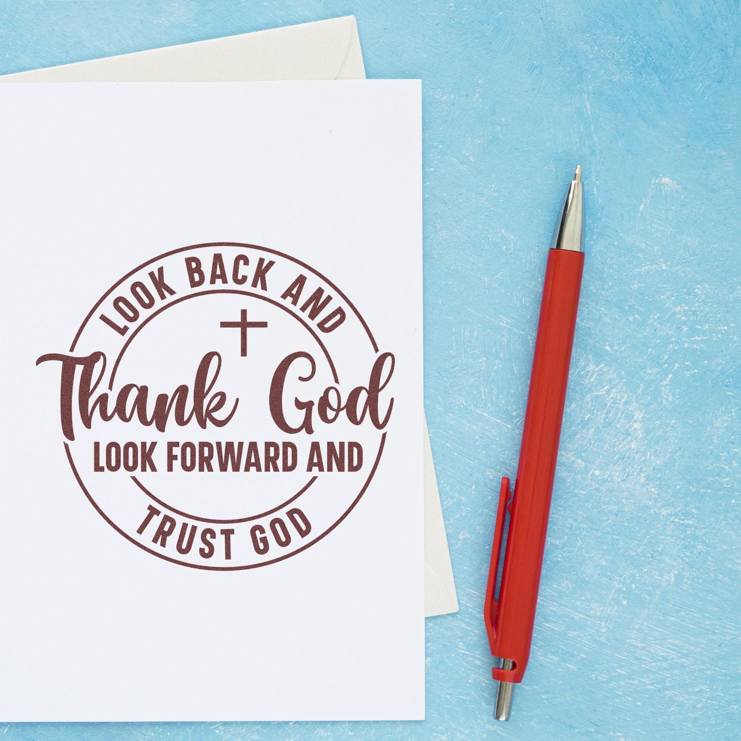 White card with brown circular faith stamp and seal reading Thank God, beside a red pen on a textured blue background.
