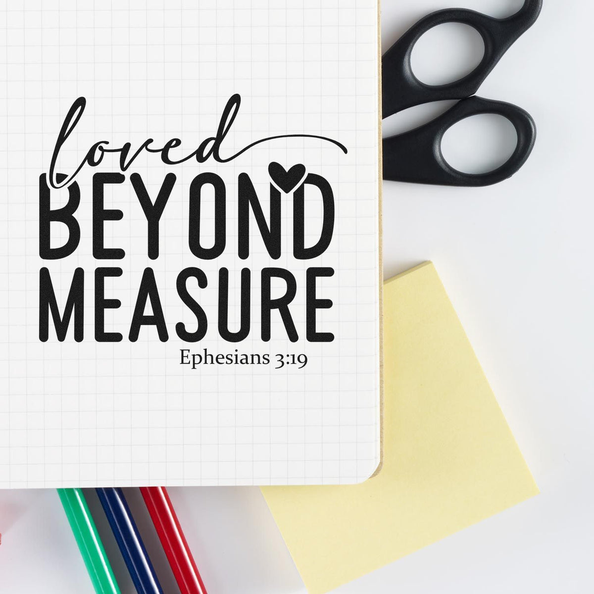Black stamp design reading loved beyond measure with Ephesians 3:19, beside black scissors, yellow note, and colored markers.