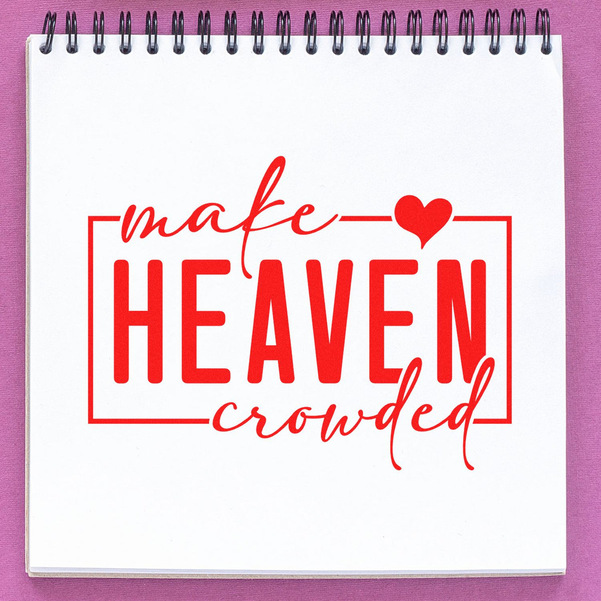 Red decorative stamp reading make HEAVEN crowded with heart on white spiral notebook, against a purple background.