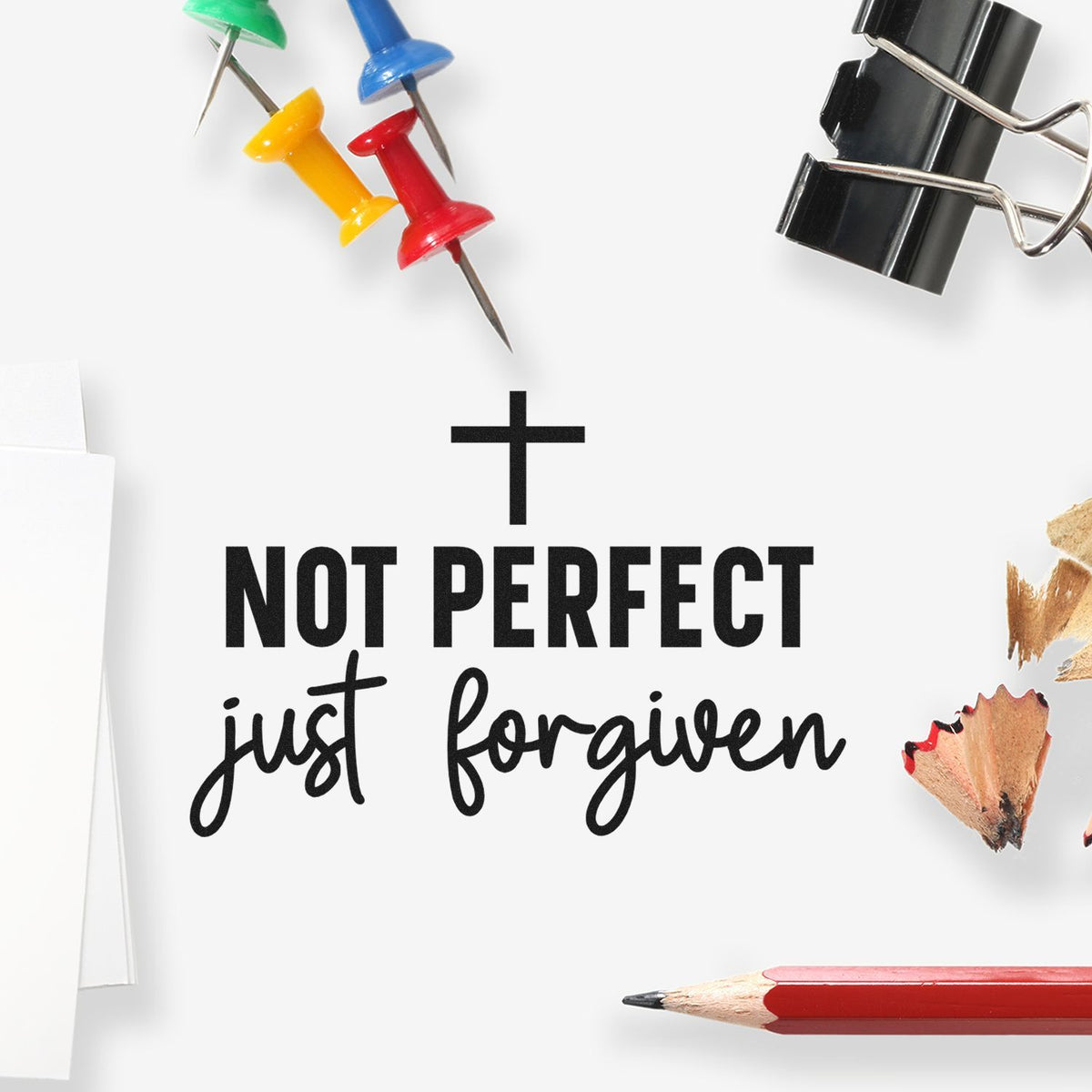 Black faith stamp design reading Not perfect, just forgiven with colorful push pins, binder clip, and red pencil.