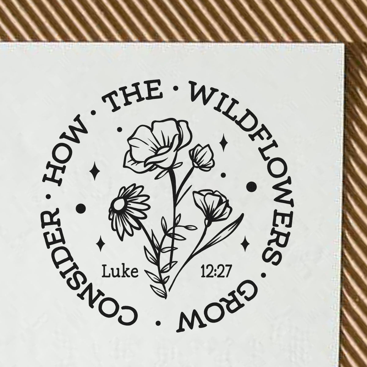 Black floral stamp seal reading Consider how the wildflowers grow with Luke 12:27 on a white background.