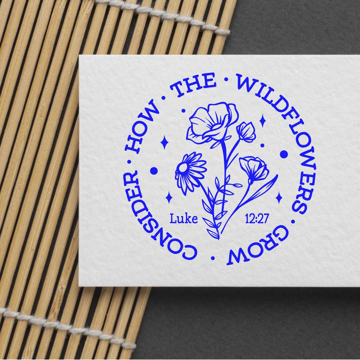 Blue floral stamp seal on white card with Consider the Wildflowers and Luke 12:27, beside a bamboo mat.