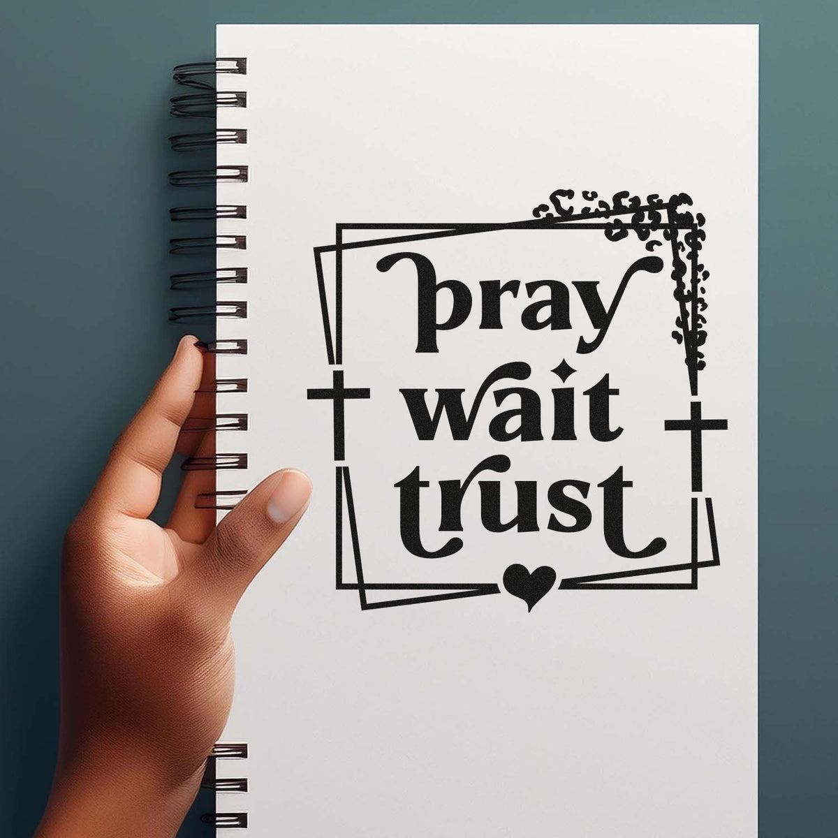 Black stamp seal design with pray wait trust, cross and heart on white spiral notebook, held by hand on teal background.