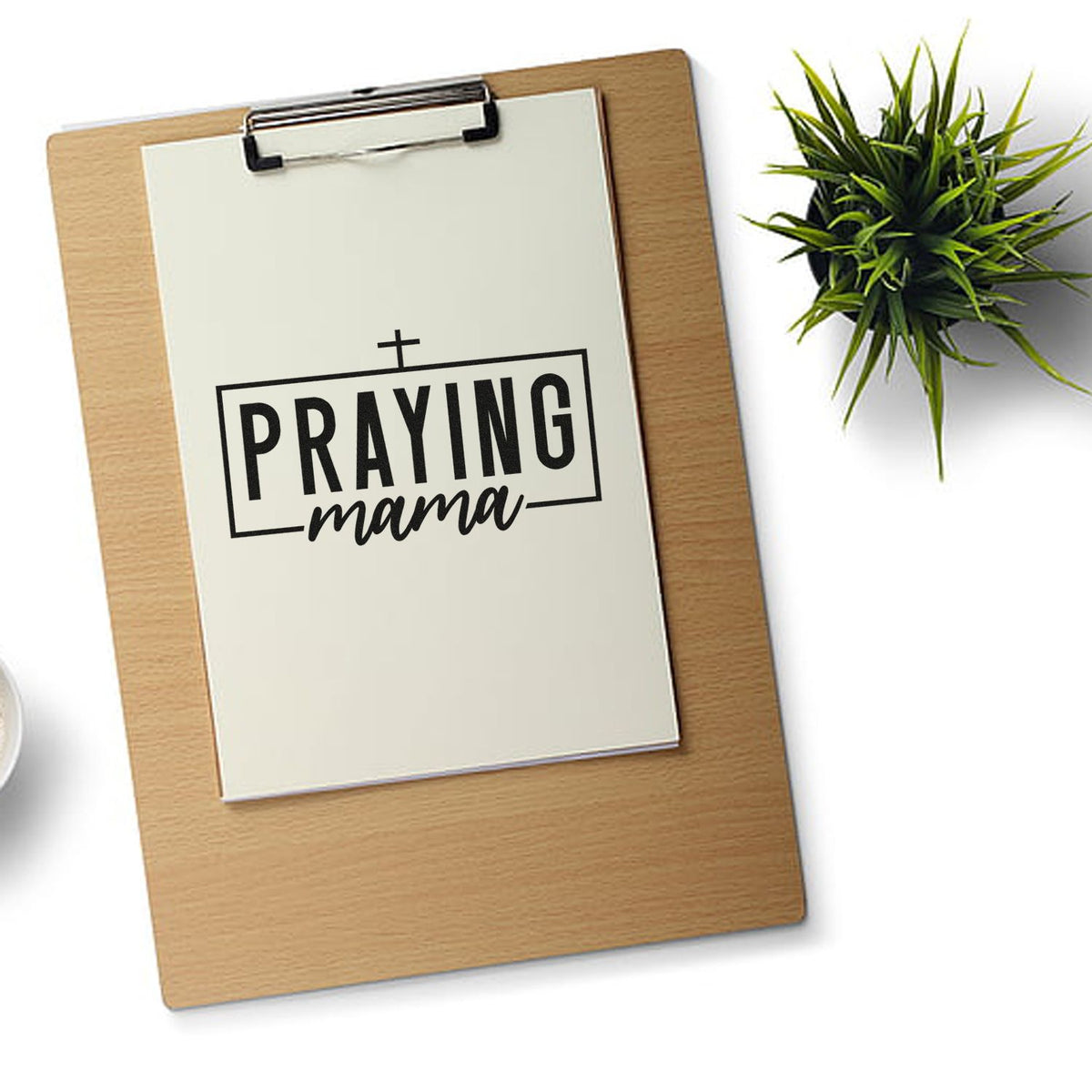 Wood clipboard with white paper stamped PRAYING mama in black, beside a green plant on a white background.