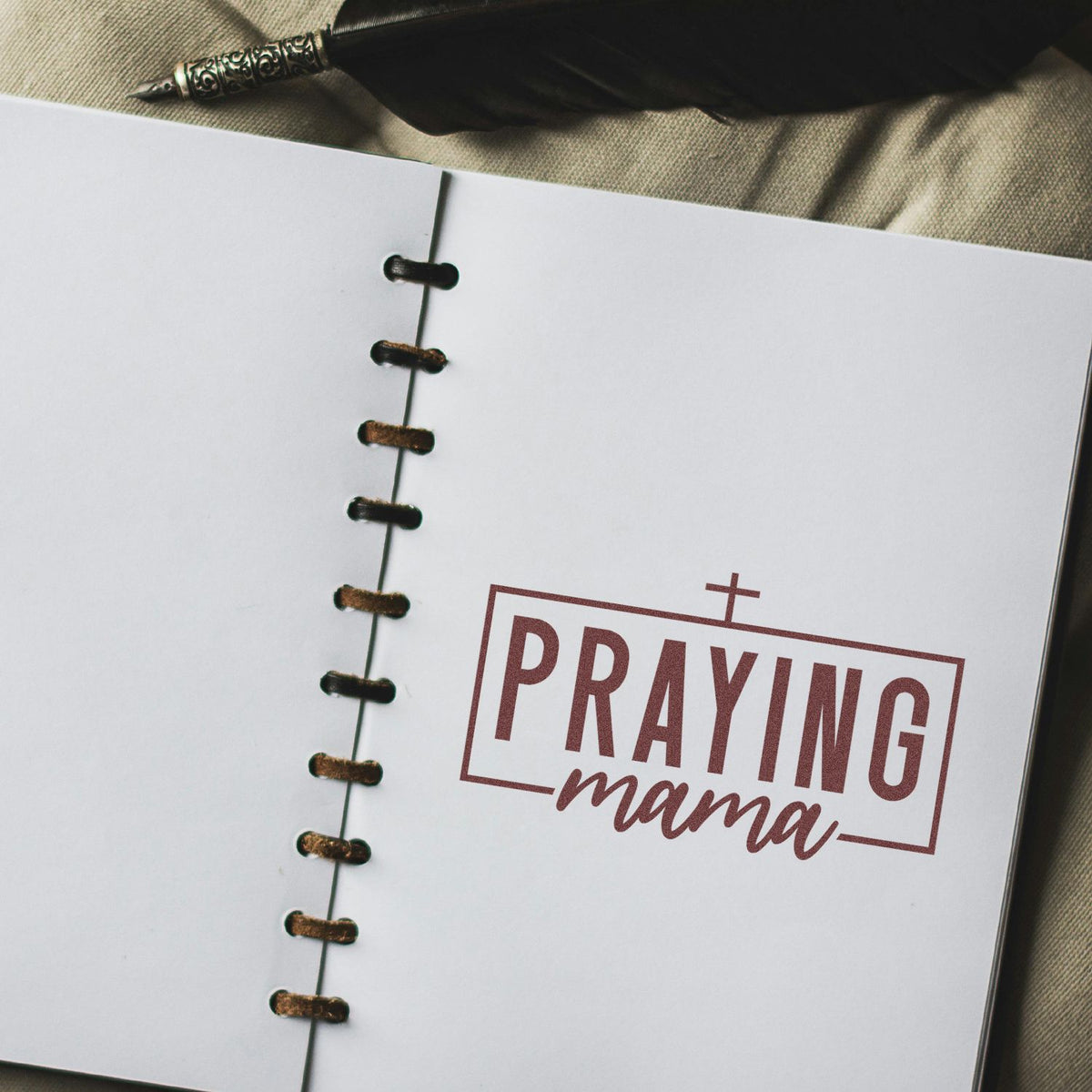 White spiral notebook with maroon PRAYING mama stamp and seal, black binding, and pen on beige fabric.