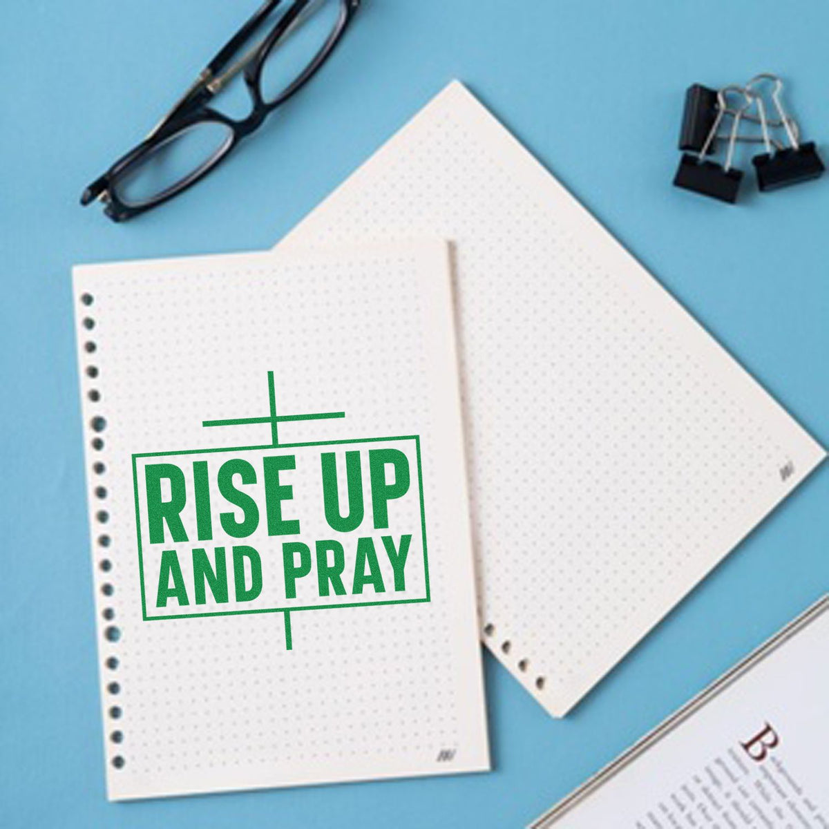 Green Rise Up and Pray stamp seal on white dotted paper, with black glasses and binder clips on a light blue desk.