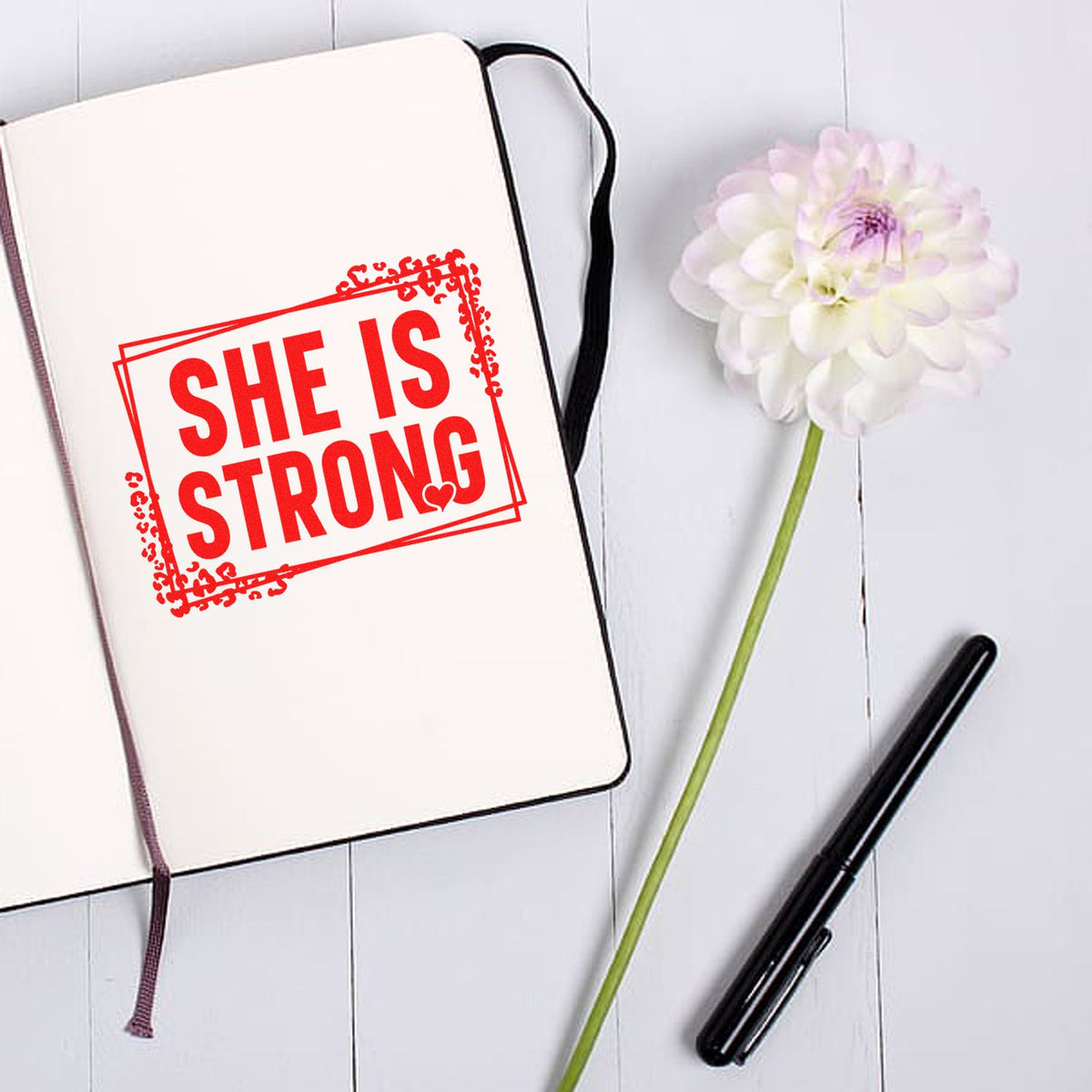 Red SHE IS STRONG stamp seal on cream notebook beside a white flower and black pen.
