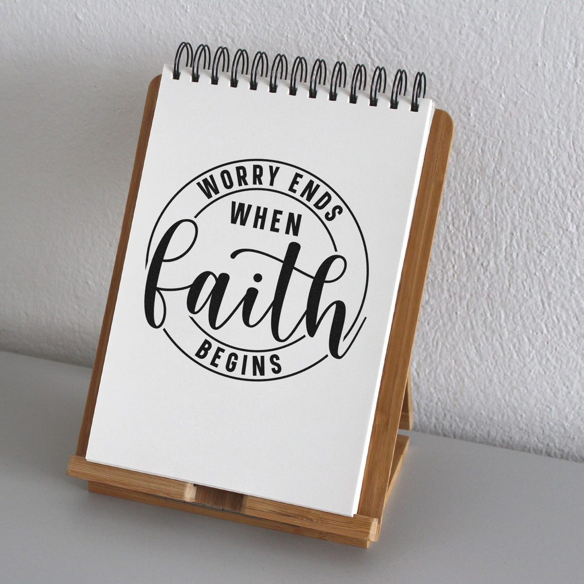 White spiral notepad on wooden stand with black faith stamp reading Worry ends when faith begins.