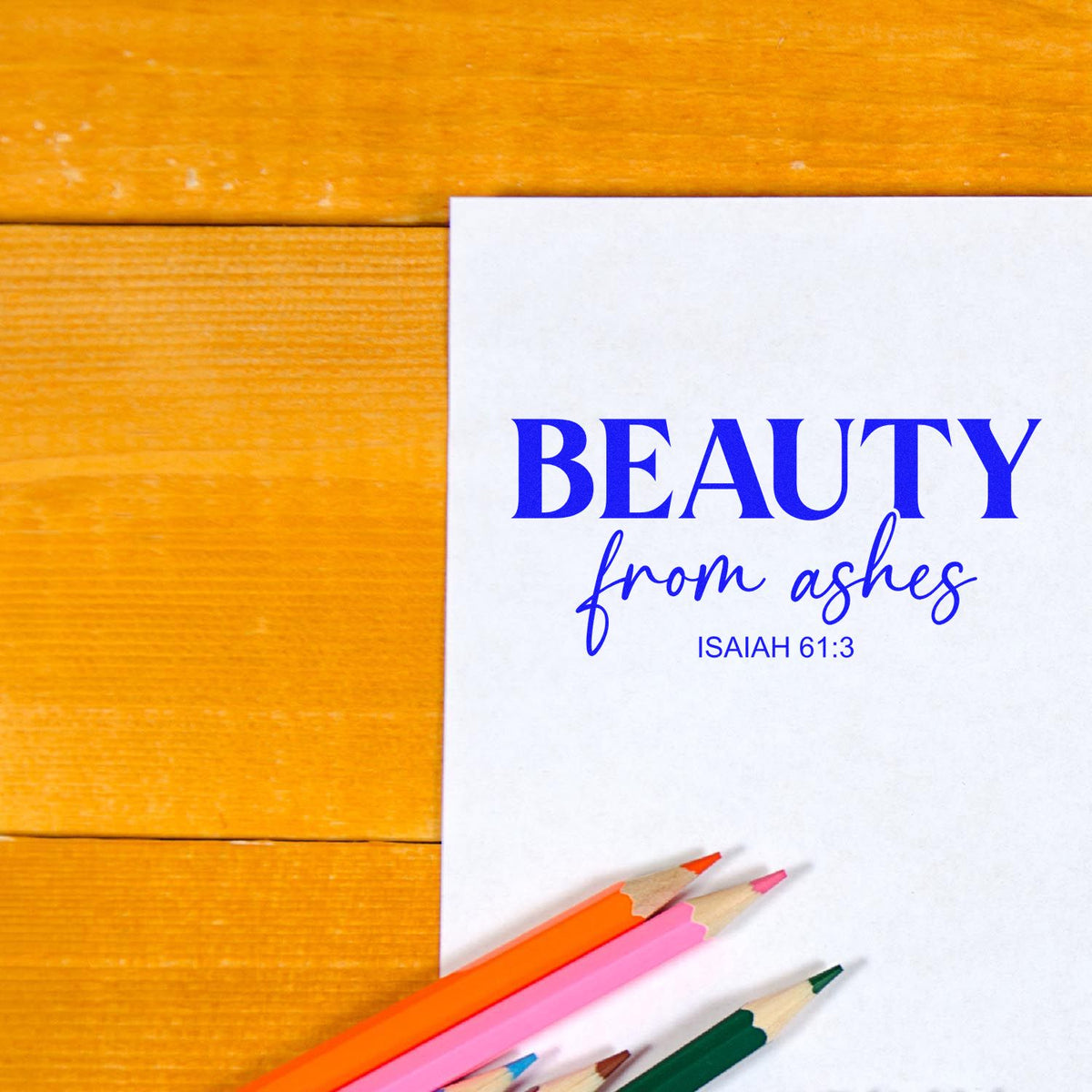 Custom stamp and seal card reading Beauty from ashes in blue, with orange, pink, and green pencils on a wood table.