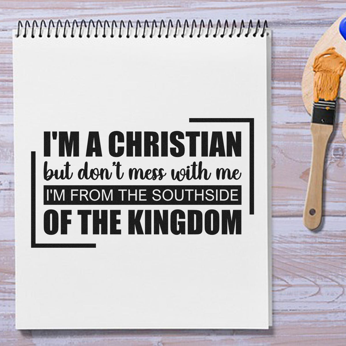 Black Christian quote on a white spiral notebook beside a tan wooden stamp and seal brush on a light wood background.