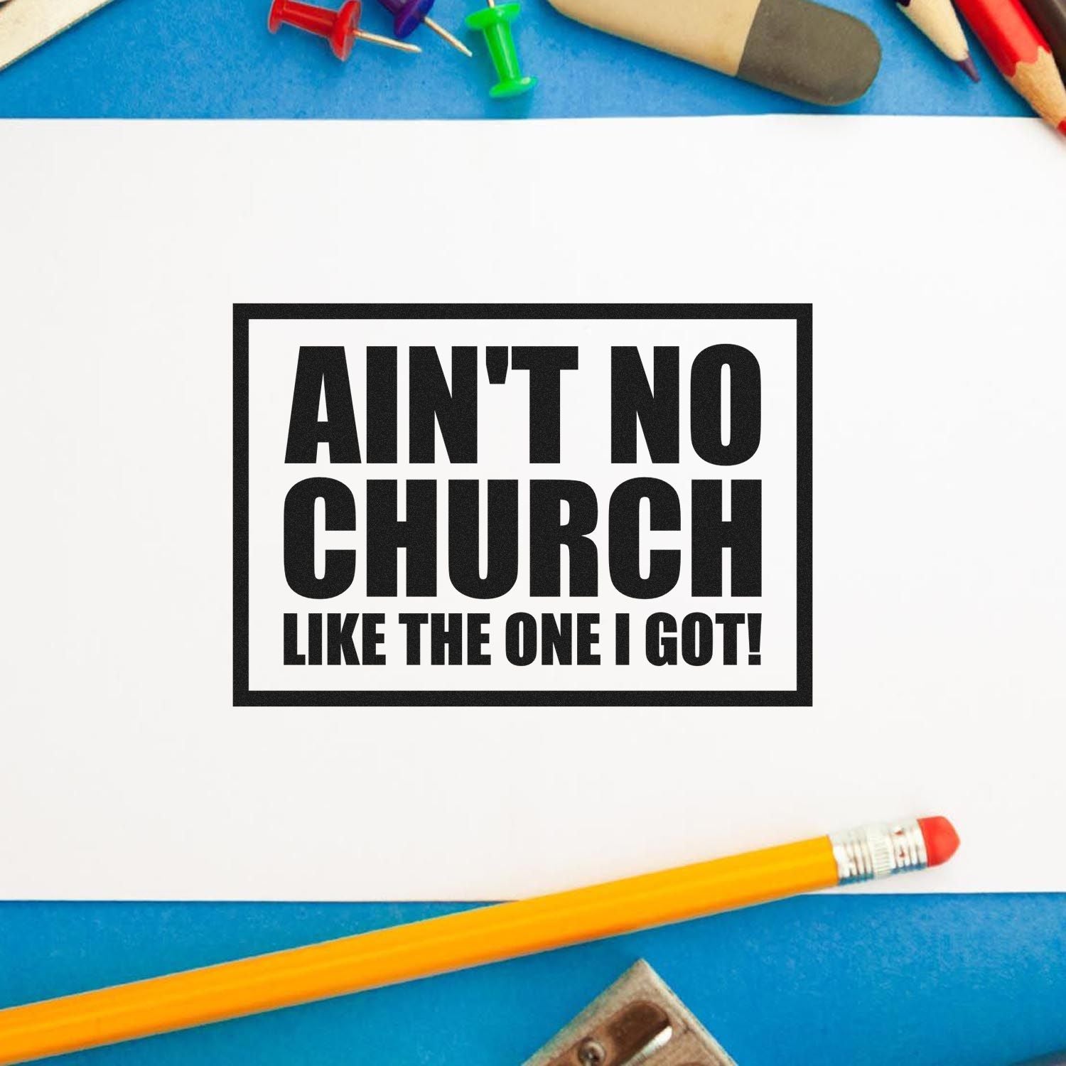 Black custom stamp/seal imprint reading AINT NO CHURCH LIKE THE ONE I GOT! on white paper, with blue background and pencils.