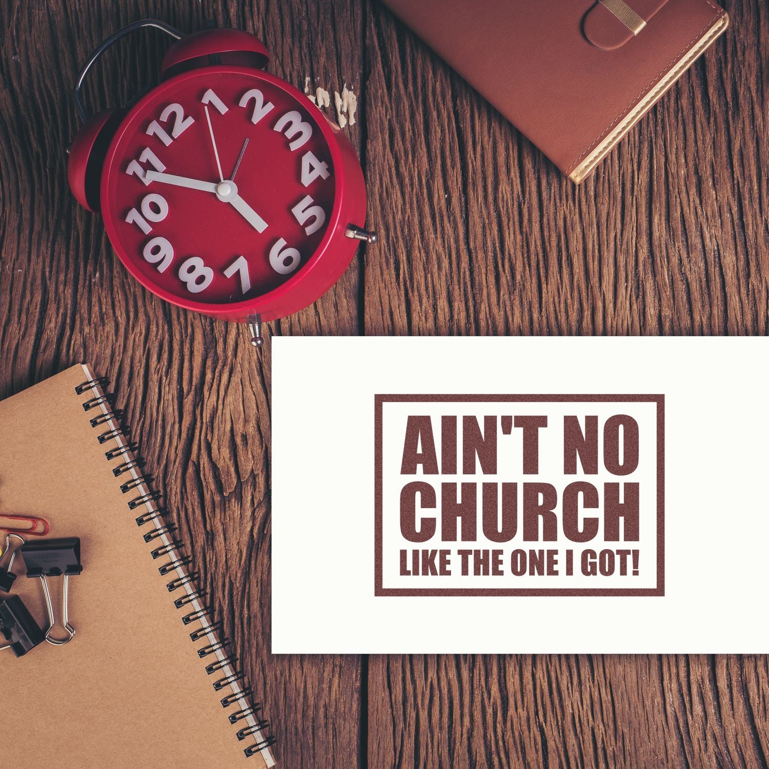 Brown stamp seal reading Aint No Church Like the One I Got! beside a red alarm clock on a rustic wood desk.