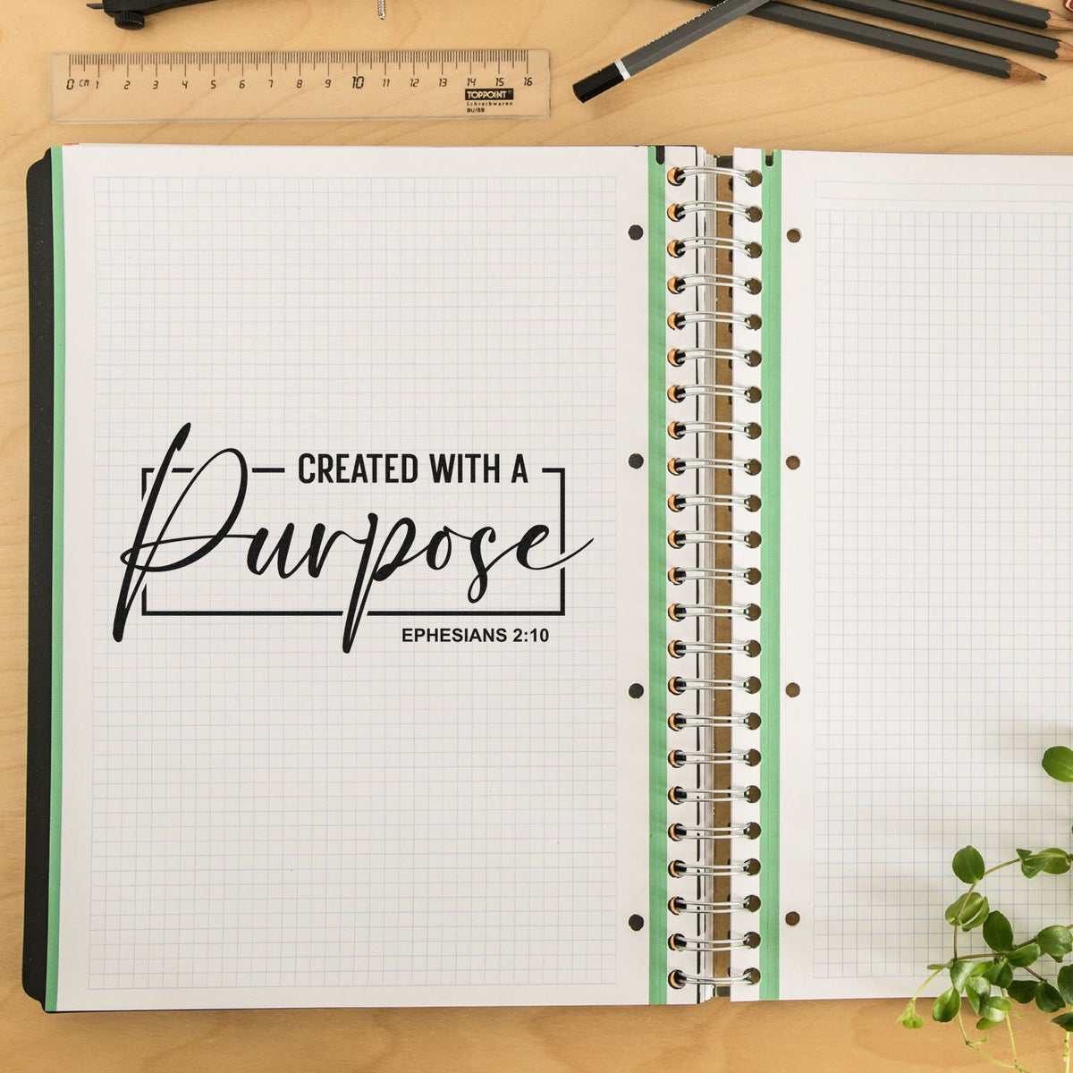 Black scripture stamp reading Created with a Purpose on white grid notebook pages, with green rings and a ruler on a wood desk.