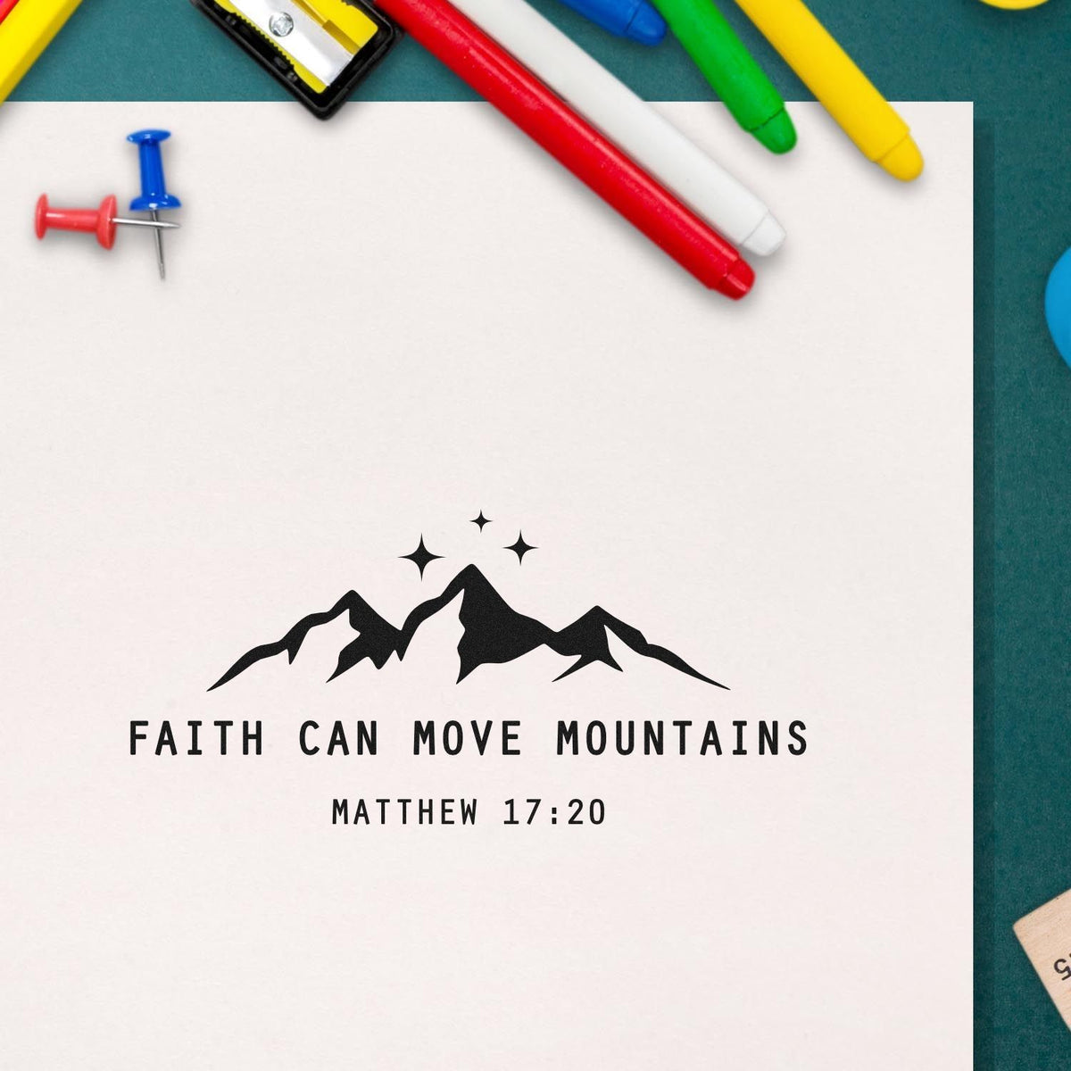 Black faith stamp seal with mountain graphic and Matthew 17:20 on white paper, surrounded by colorful pens and push pins.
