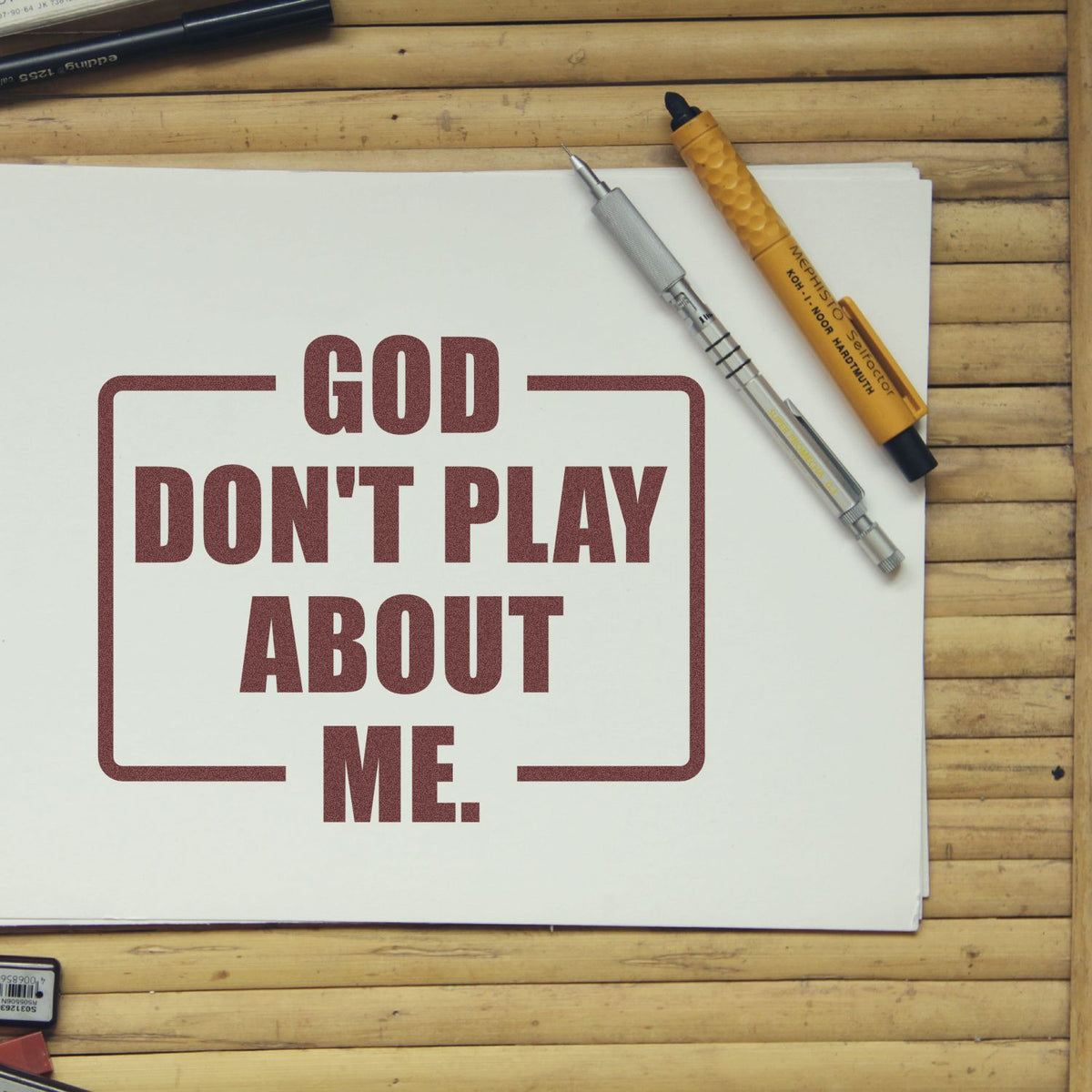Maroon rubber stamp-style text GOD DONT PLAY ABOUT ME on white paper, with stamp and seal tools on a wooden desk.