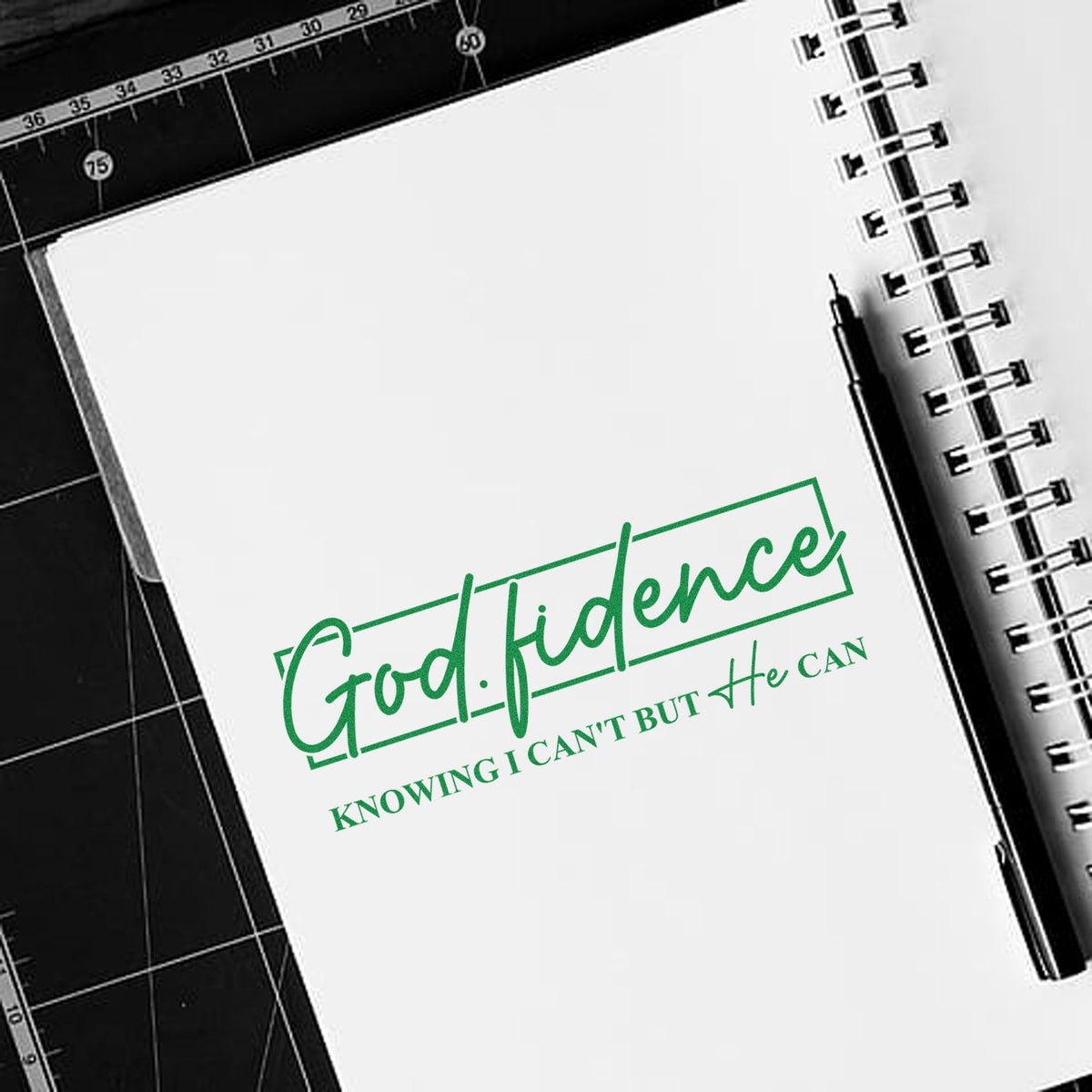 Green God-fidence custom stamp seal on a white notebook, with black pen, spiral binding, and black cutting mat.