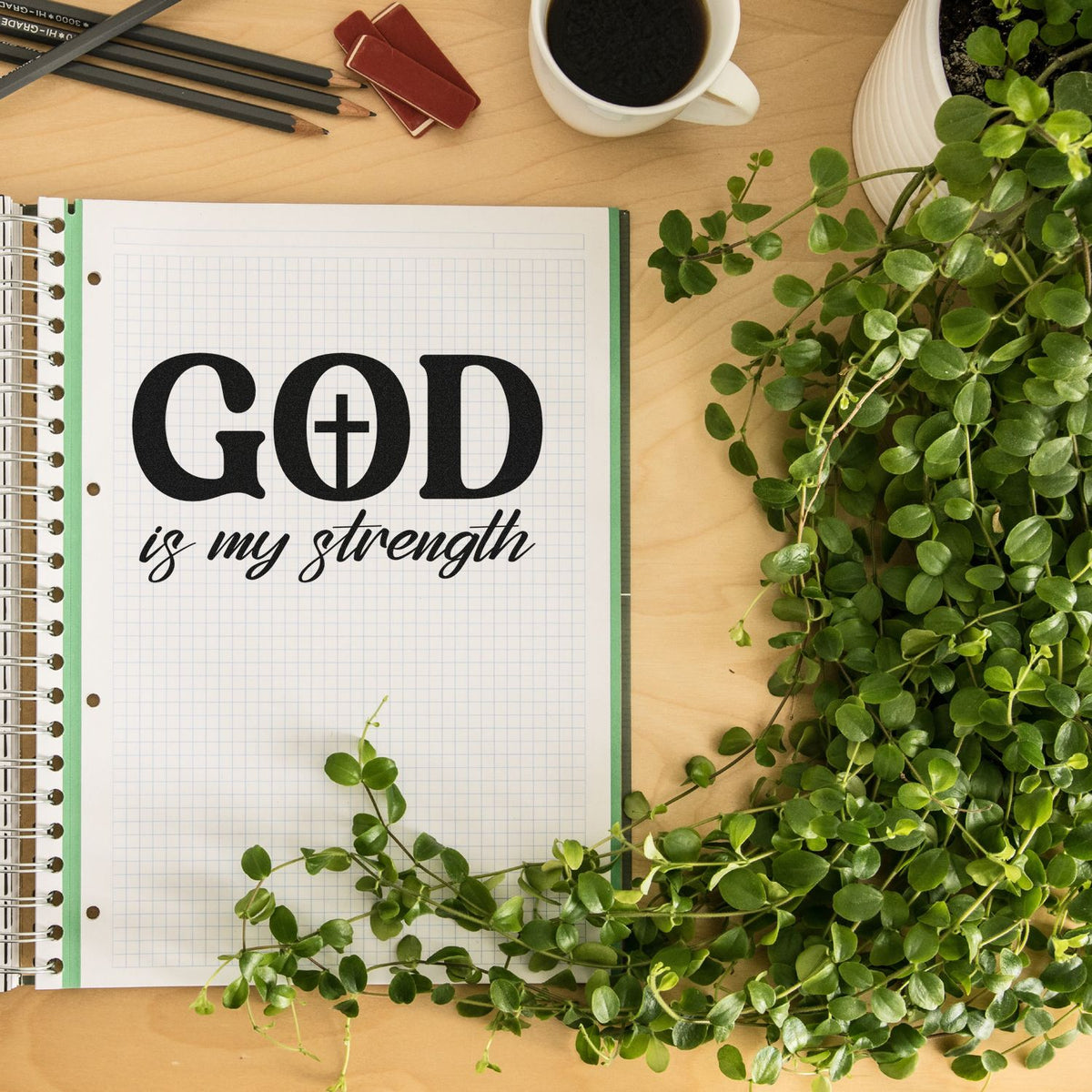 Black stamp and seal on graph notebook reading GOD is my strength, with green leaves, pencils, coffee, and a wood desk.