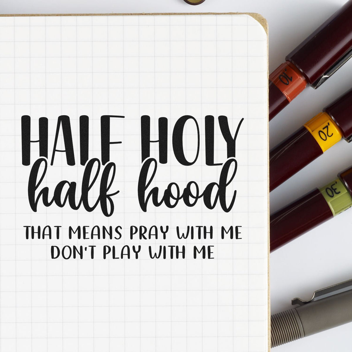 White grid notebook page with black Half Holy Half Hood stamp, beside burgundy stamp and seal tools in orange, yellow, and olive.