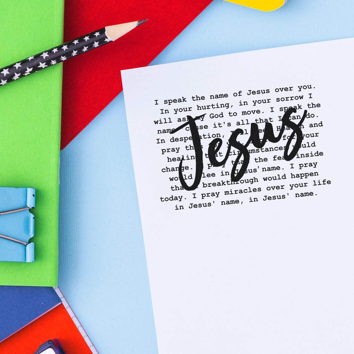 Black 'Jesus' stamp on white prayer card, with colorful stationery, star pencil, and binder clip.
