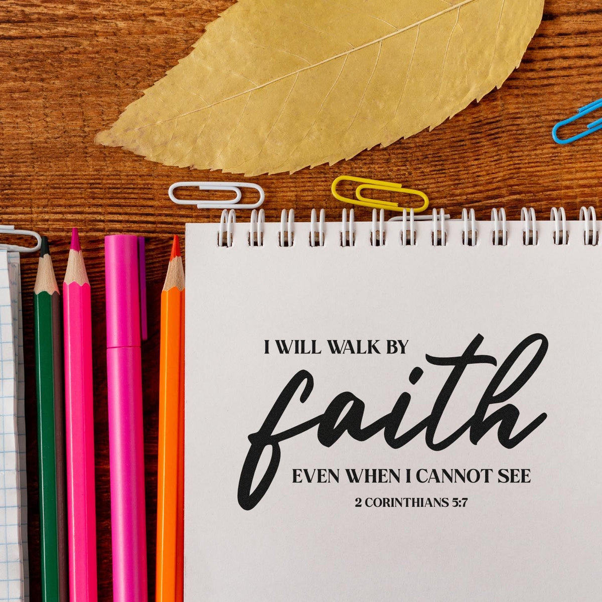 Faith stamp and seal stationery flat lay with white notebook, black script quote, colorful pencils, and tan leaf.