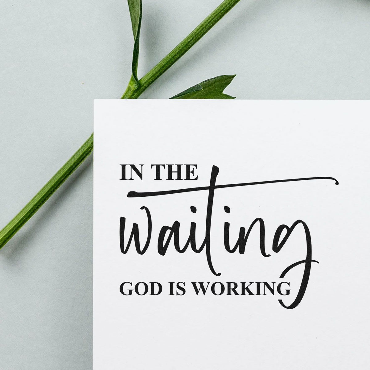 Black faith quote stamp seal on white card with green stem: In the waiting, God is working.