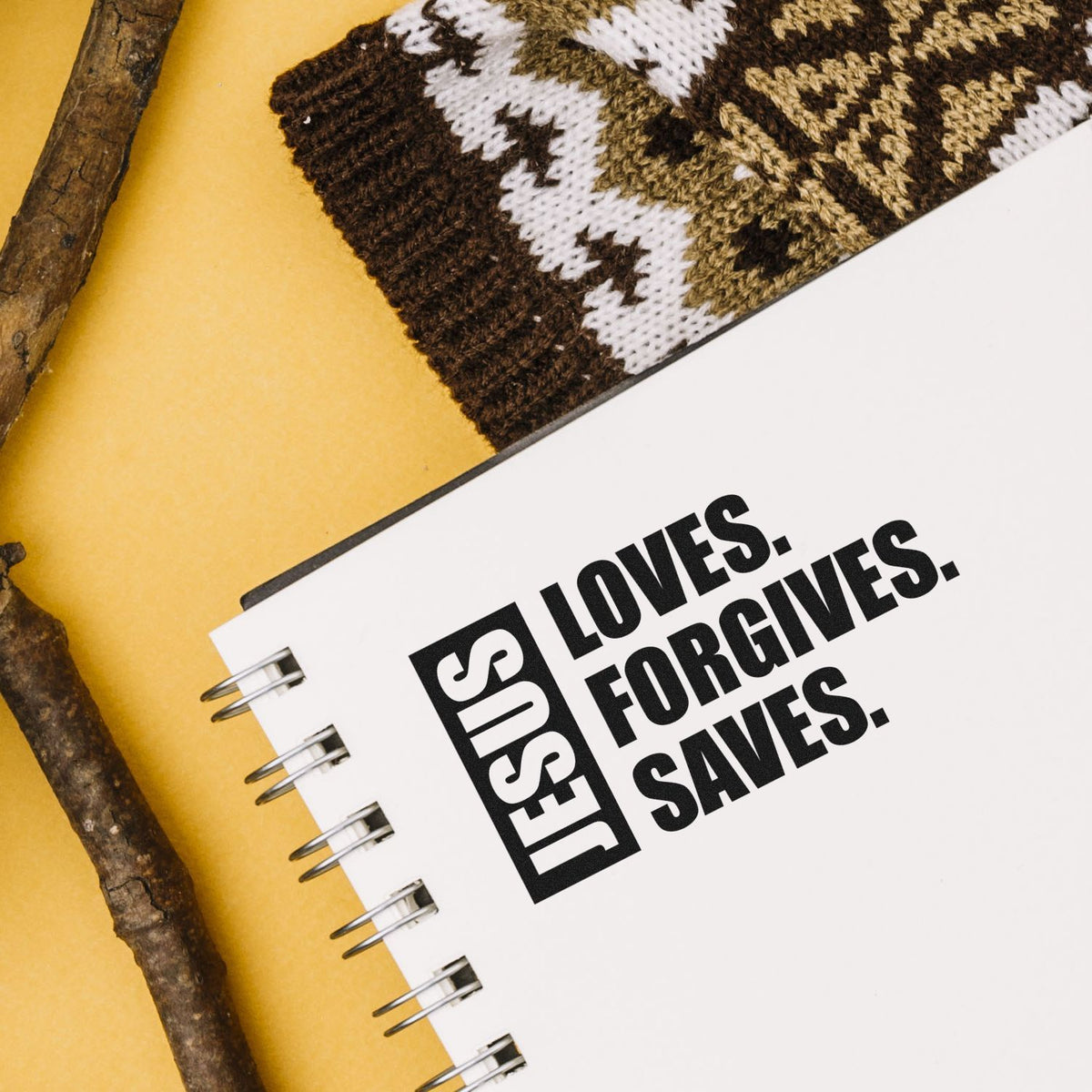 Black custom stamp and seal on white spiral notebook, with bold Jesus loves. Forgives. Saves. on yellow background.