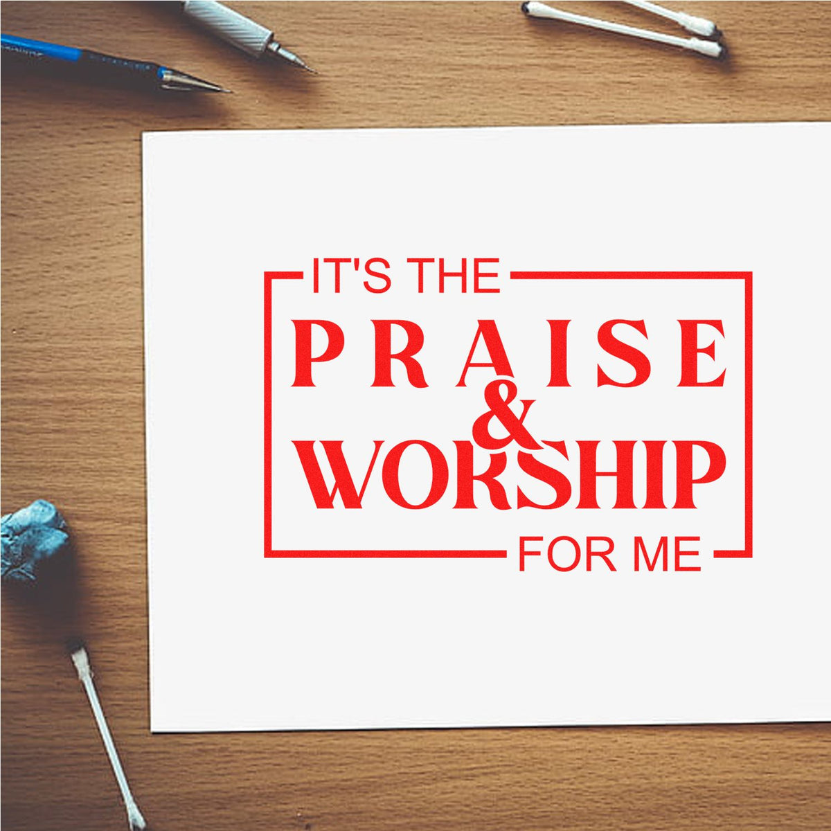 Red praise and worship stamp/seal design on white paper, framed in red on a wood desk with art tools
