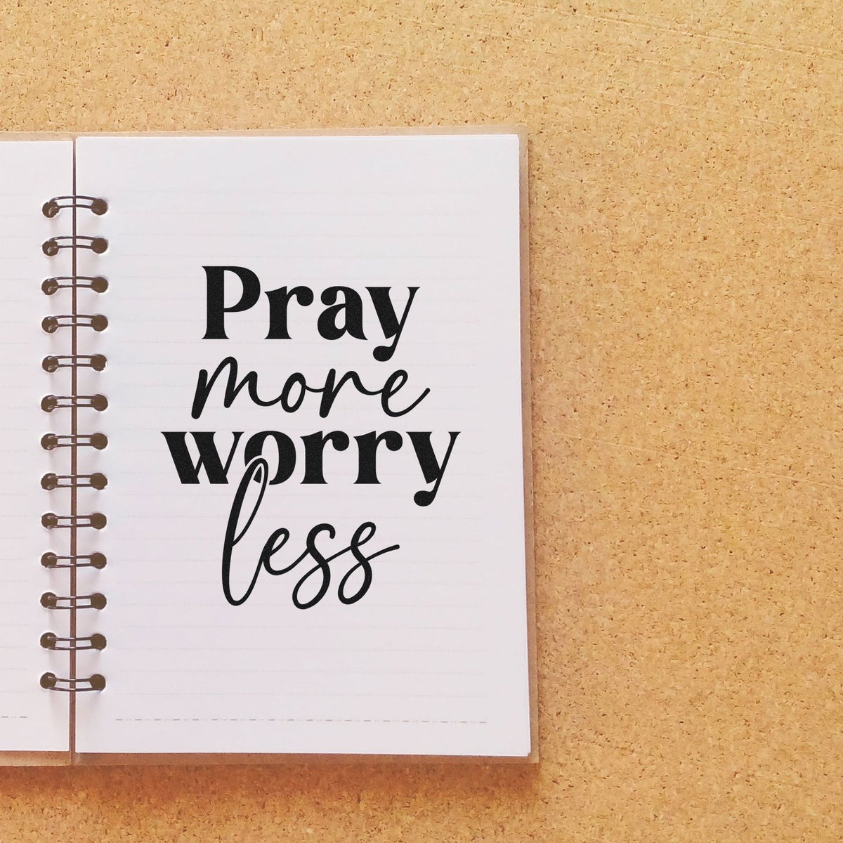 Black Pray more worry less stamp/seal on a white spiral notebook over a tan cork background.