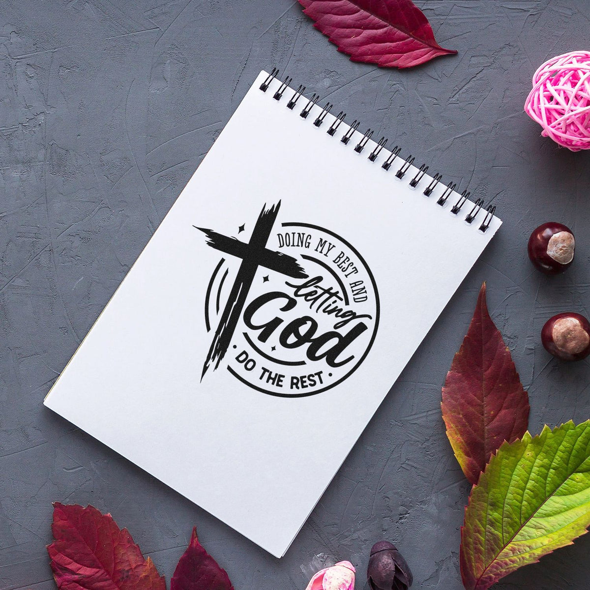 Black custom stamp/seal with cross and faith quote on white spiral notebook, set on gray textured surface with red leaves.