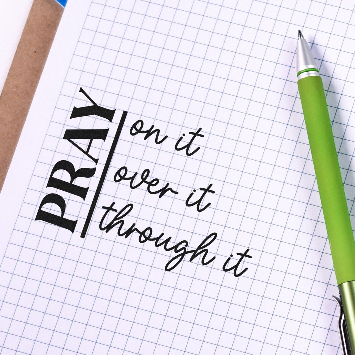 Black custom stamp seal reading Pray on it, over it, through it on graph paper, with a green pen at right.