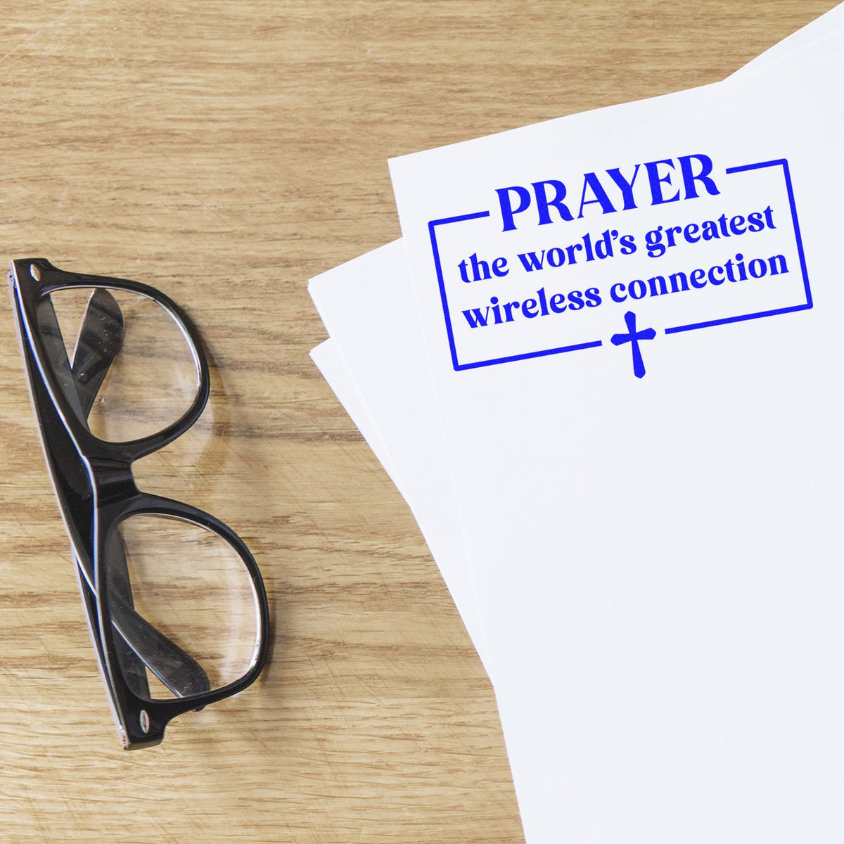 Blue stamp-and-seal print reading PRAYER... on white paper beside black eyeglasses on a wood desk