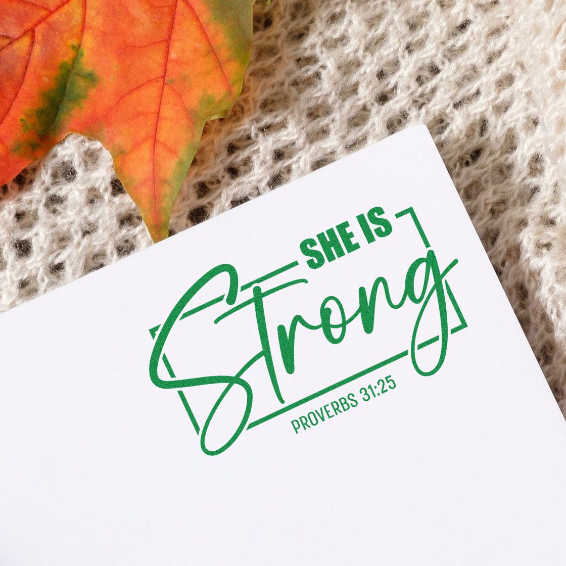 Green custom stamp and seal reading She Is Strong with Proverbs 31:25 on white paper beside an autumn leaf.