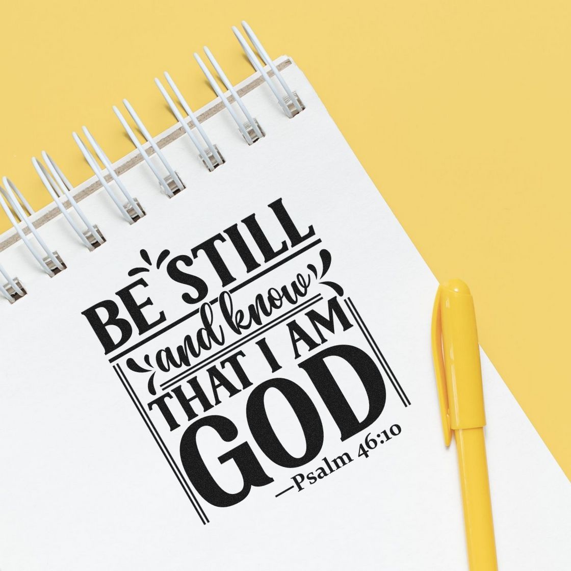 White spiral notebook with black stamp-seal scripture design, beside a yellow pen on a yellow background.